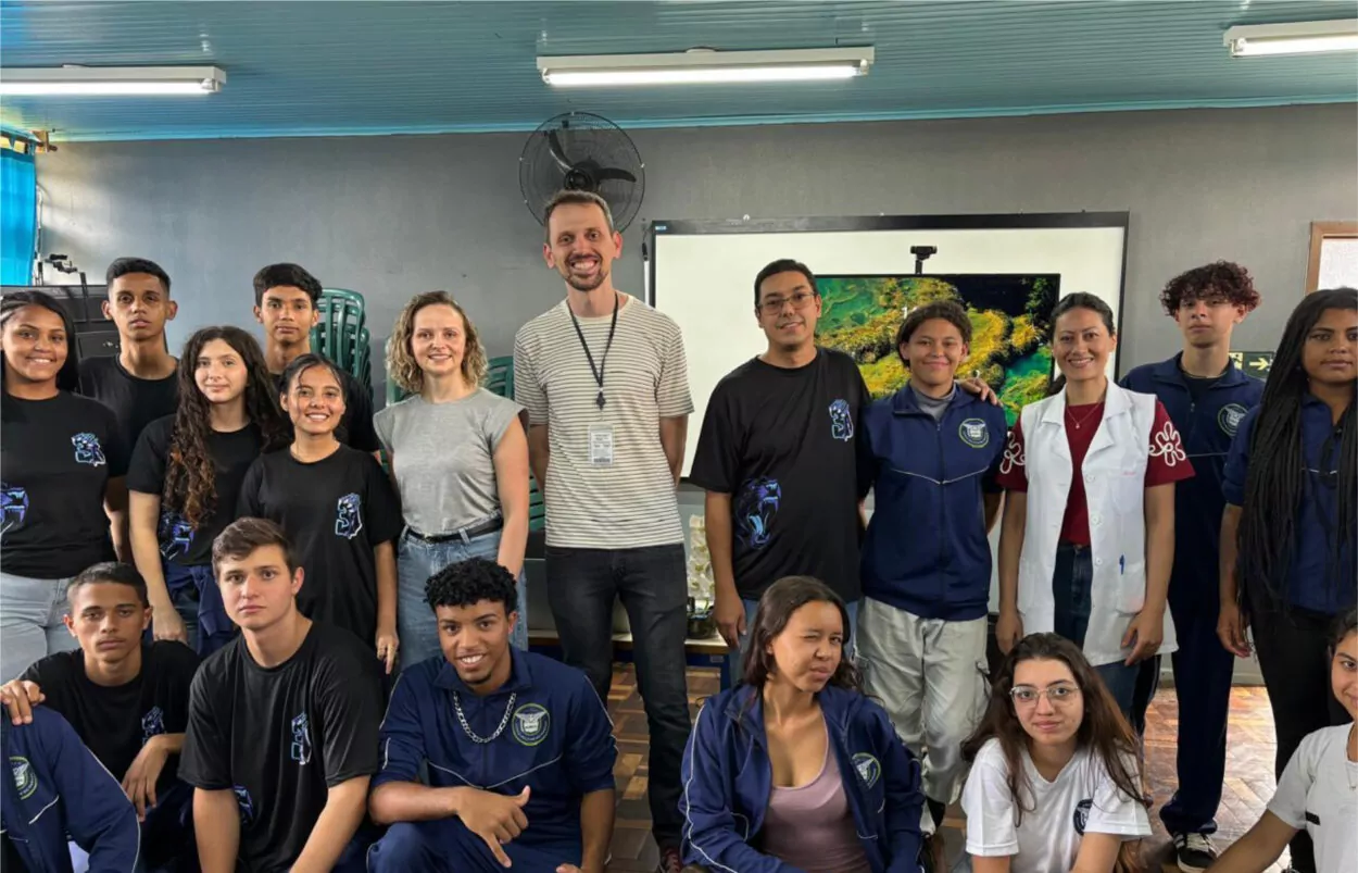 A imagem mostra um grupo de pessoas em uma sala de aula. O grupo é composto por estudantes e professores, alguns vestindo uniformes escolares e outros com roupas casuais. Há uma tela ao fundo exibindo uma imagem da natureza, possivelmente um ambiente subaquático ou florestal. O ambiente tem um teto azul com luminárias e ventiladores. Algumas pessoas estão sorrindo, enquanto outras mantêm expressões neutras.