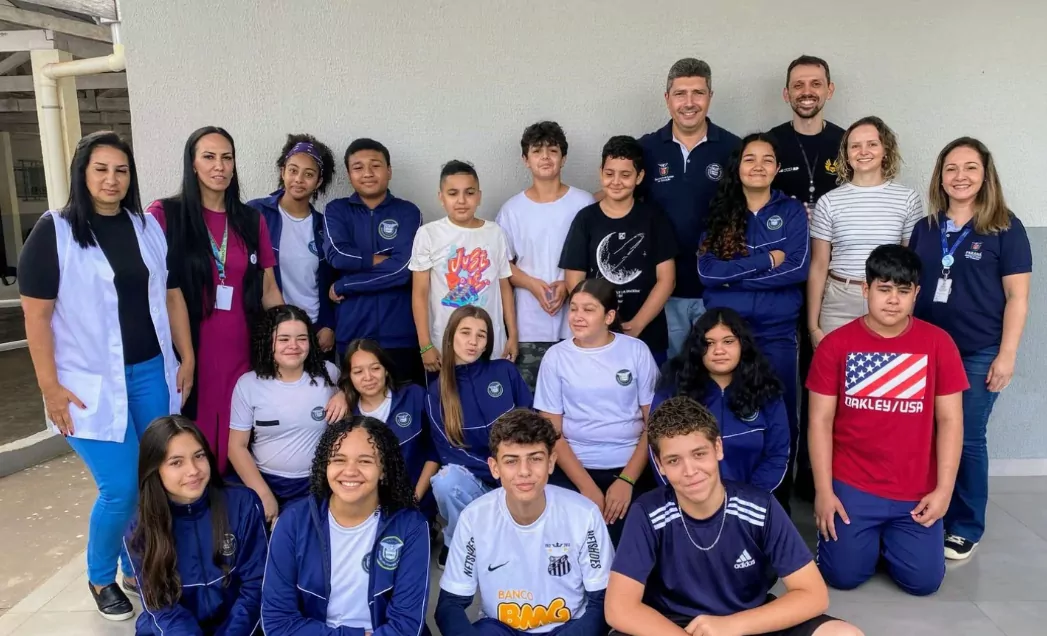 A imagem mostra um grupo de pessoas posando para uma foto em frente a uma parede pintada de branco, em ambiente escolar. O grupo é composto por adolescentes e adultos, alunos e professores. Eles estão sorrindo e parecem felizes e animados. Os adolescentes estão usando uniformes escolares azuis e brancos, enquanto os adultos estão vestidos de forma casual. A parede branca atrás deles tem um padrão de grade cinza claro.