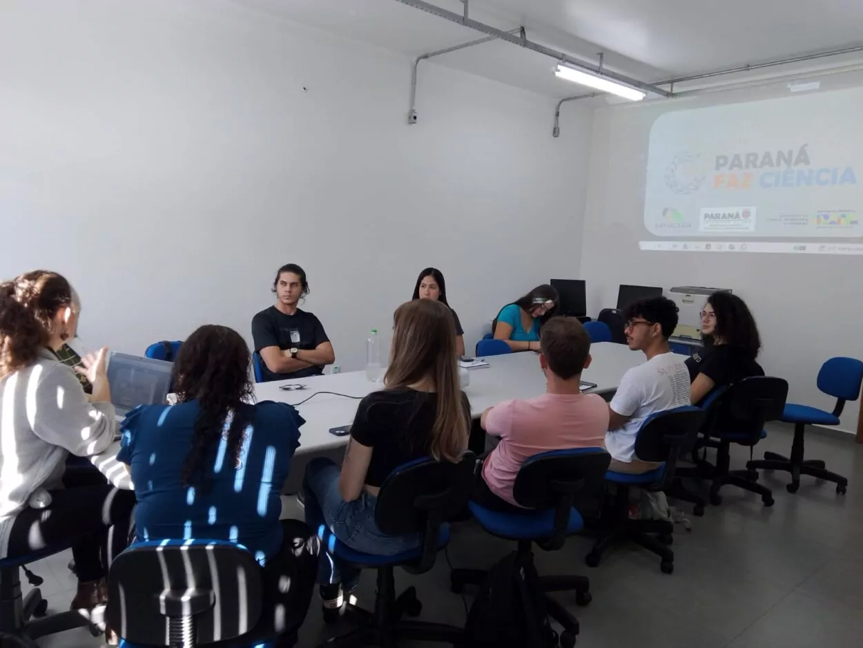A foto mostra a equipe da UEPG reunida ao redor de uma mesa em uma sala. Uma apresentação do projeto "Paraná Faz Ciência" é projetada na parede ao fundo. Uma pessoa, posicionada à esquerda da imagem, utiliza um notebook enquanto conversa com o grupo. Outras pessoas acompanham a fala, com anotações e materiais sobre a mesa. O espaço conta com iluminação natural e artificial.