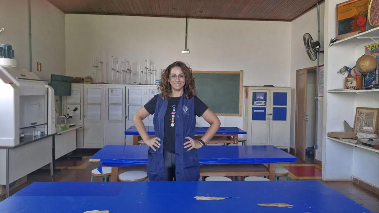 A imagem mostra a professora Eglaia Cheron, coordenadora do clube “Discovery Kennedy’s”, no laboratório de ciências do Colégio Estadual Cívico-Militar Presidente Kennedy, em Rolândia. A docente está no centro da foto, atrás de uma mesa azul. Em volta, há prateleiras brancas com objetivos utilizados no meio científico e outros do cotidiano, como um globo terrestre. No fundo, há ainda um quadro negro e outras mesas azuis.