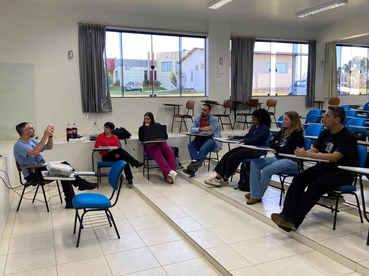 Fotografia de uma sala de aula com janelas grandes ao fundo, por onde entra luz natural. Um grupo de sete pessoas está reunido, sentadas em carteiras escolares organizadas em semicírculo. À esquerda, um homem de barba, usando camiseta azul, está sentado e gesticulando enquanto fala. À sua frente, os outros participantes — três mulheres e três homens — prestam atenção, alguns com cadernos ou notebooks no colo. Há duas garrafas de refrigerante e alguns lanches sobre uma cadeira próxima à parede. A sala é clara, com piso branco e cadeiras azuis. Ao fundo, é possível ver o pátio externo e alguns prédios.