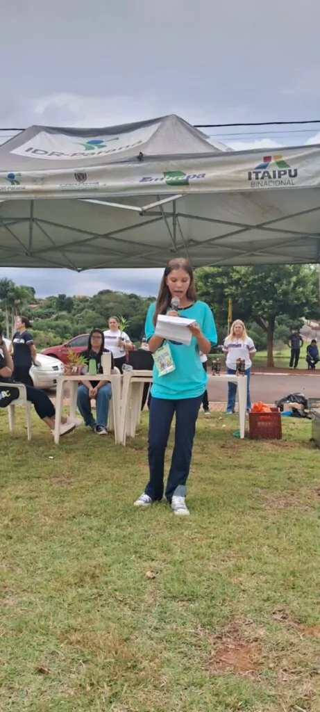 A imagem mostra uma menina em pé, em um espaço ao ar livre, lendo um texto em voz alta com a ajuda de um microfone. Ela está segurando algumas folhas de papel e parece estar concentrada na leitura. A menina veste camiseta azul e calça escura, com um crachá pendurado no pescoço. Ao fundo, há uma tenda com logotipos de instituições como IDR-Paraná, Embrapa, Itaipu Binacional e o brasão do município de Borrazópolis, indicando a parceria entre diferentes órgãos em um evento educacional e ambiental. Atrás dela, algumas pessoas estão sentadas e observando atentamente, entre elas adultos e estudantes. Também é possível ver mesas plásticas com vasos de plantas, reforçando o tema ambiental, e ao fundo, carros e árvores, indicando que o evento ocorre em uma área aberta urbana, próxima a escola. A cena transmite a ideia de um momento importante de participação estudantil pelo meio ambiente, educação e cidadania.