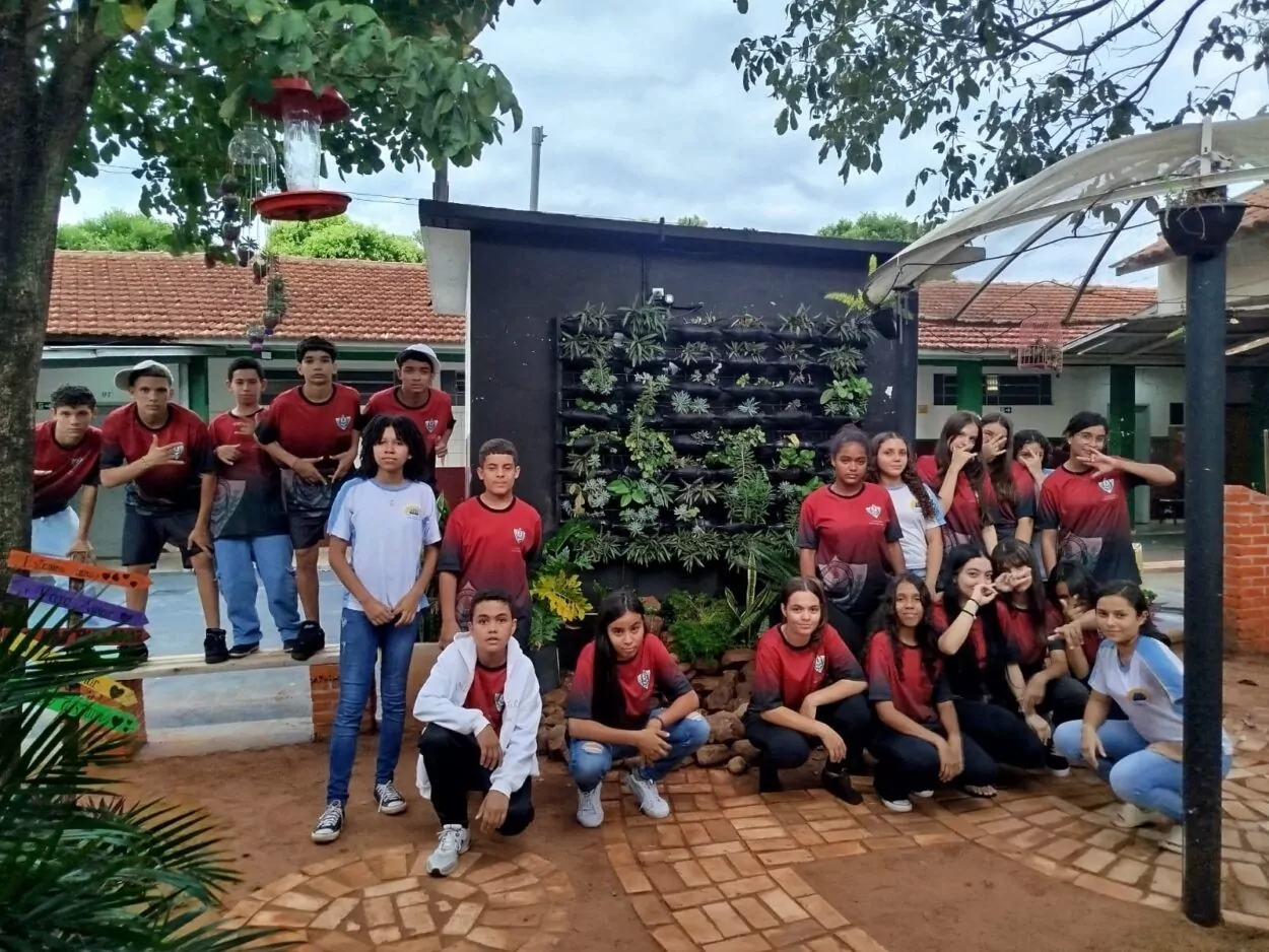 A foto é do Clube de Ciências Paisagismo Sustentável. A imagem mostra um grupo de estudantes posando em frente a um jardim vertical instalado numa parede preta, no que parece ser o pátio da escola. A maioria dos alunos estão usando camisetas vermelhas com o logotipo do colégio. O jardim vertical está bem organizado, com diversas plantas ornamentais, dando um aspecto verde e ecológico ao espaço. À esquerda, vê-se uma árvore com comedouros para beija-flores pendurados, e, à direita, uma estrutura com cobertura translúcida. O chão está revestido com blocos de pedra formando um caminho decorativo. O ambiente transmite uma sensação de envolvimento com a natureza, sendo resultado de um projeto ligado ao Clube Paisagismo Sustentável.