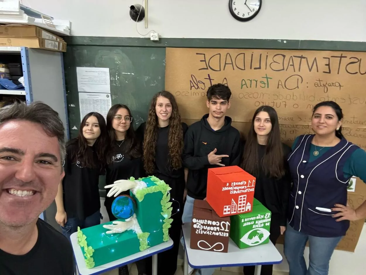 A foto é do Clube de Ciências “Planeta Consciente Paraná Faz Ciência Maker”. A imagem mostra um grupo de sete pessoas (cinco jovens e dois adultos) posando sorridentes em uma sala de aula. Todos parecem estar participando de um projeto escolar relacionado à sustentabilidade. Atrás do grupo, há um cartaz colado na lousa verde, com o título “SUSTENTABILIDADE” e outros textos escritos à mão, mencionando “Projeto: DA TERRA À TELA” e palavras como “futuro” e “sustentável”. Na frente do grupo, há duas mesas com maquetes. Uma delas exibe mãos segurando um globo terrestre, cercado por folhas verdes artificiais e luzes decorativas. A outra tem cubos coloridos com ícones e textos representando os Objetivos de Desenvolvimento Sustentável (ODS), como o ODS 11 (Cidade e Comunidade Sustentável), ODS 12 (Consumo e Produção Responsáveis) e o ODS 13 (Ação Contra a Mudança Global do Clima). O ambiente transmite um clima de cooperação e engajamento estudantil.