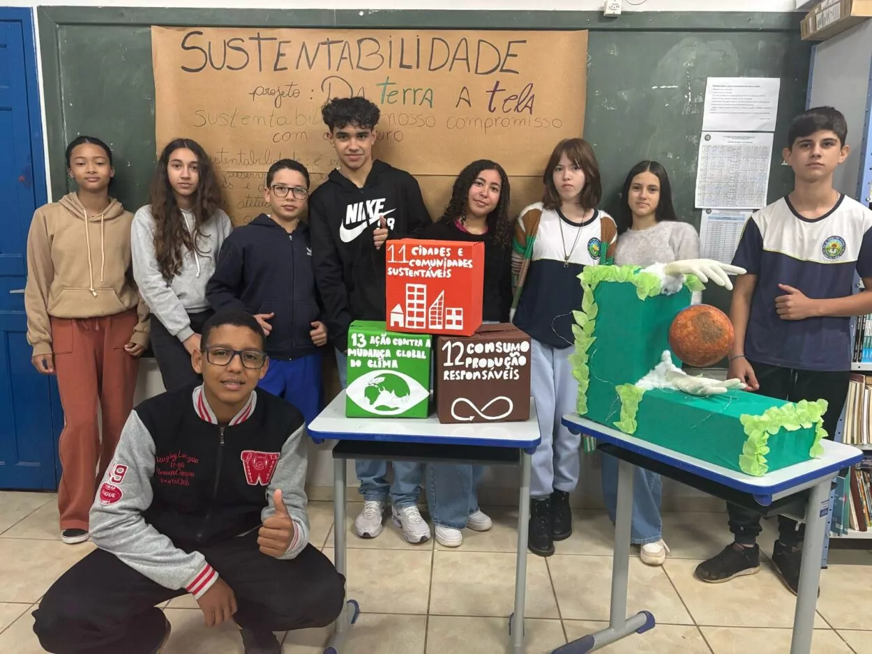 A imagem mostra um grupo de dez estudantes posando em sala de aula, diante de um mural com o tema "SUSTENTABILIDADE", escrito em um cartaz de papel fixado na lousa. O cartaz traz o título do projeto “Da Terra à Tela” e o subtítulo "Sustentabilidade: nosso compromisso com o futuro". Na frente do grupo, há duas mesas com maquetes. Na mesa central, há três cubos grandes que representam Objetivos de Desenvolvimento Sustentável (ODS): ODS 11: "Cidades e comunidades sustentáveis" (cubo vermelho), ODS 12: "Consumo e produção responsáveis" (cubo marrom) e ODS 13: "Ação contra a mudança global do clima" (cubo verde). Na mesa à direita, está uma escultura artística com uma estrutura verde em forma de “L”, decorada com folhas artificiais e luzes. Duas mãos seguram o planeta Terra em estado de degradação. Os estudantes demonstram engajamento com o tema.