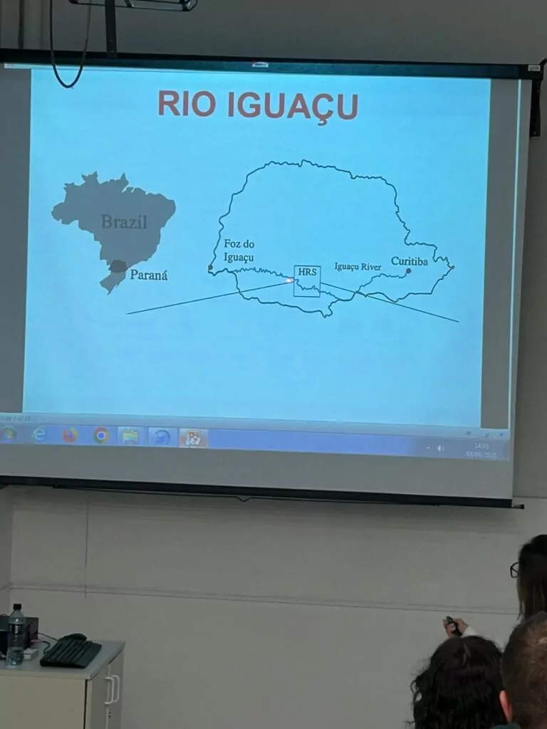 A galeria de fotos mostra momentos da visita dos clubistas à UFPR. Trata-se de ambientes fechados como um auditório e um laboratório. Há fotos da palestra que mostra várias cadeiras enfileiradas ocupadas por estudantes e professores, que estão assistindo à palestrante e à projeção de slide sobre o Rio iguaçu. Nas fotos do laboratório, mostra pequenos grupos de estudantes ouvindo orientações dos professores, usando microscópios e tablets, fazendo anotações. Todos estão vestindo jalecos com expressões de concentração e interesse.