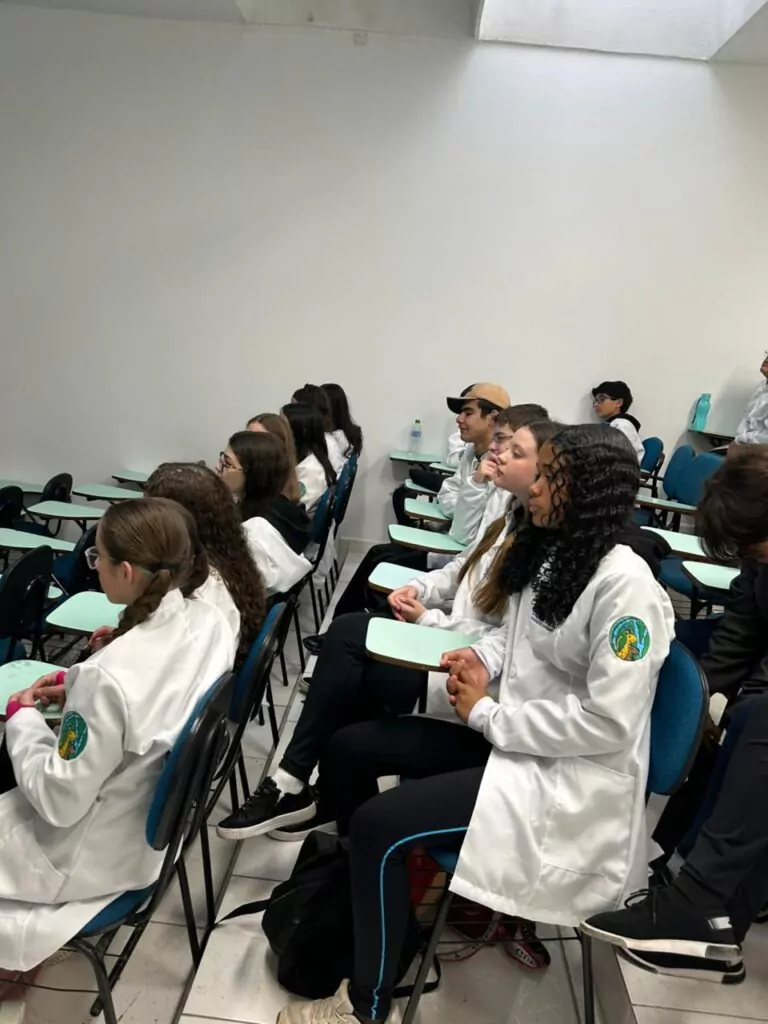 A galeria de fotos mostra momentos da visita dos clubistas à UFPR. Trata-se de ambientes fechados como um auditório e um laboratório. Há fotos da palestra que mostra várias cadeiras enfileiradas ocupadas por estudantes e professores, que estão assistindo à palestrante e à projeção de slide sobre o Rio iguaçu. Nas fotos do laboratório, mostra pequenos grupos de estudantes ouvindo orientações dos professores, usando microscópios e tablets, fazendo anotações. Todos estão vestindo jalecos com expressões de concentração e interesse.