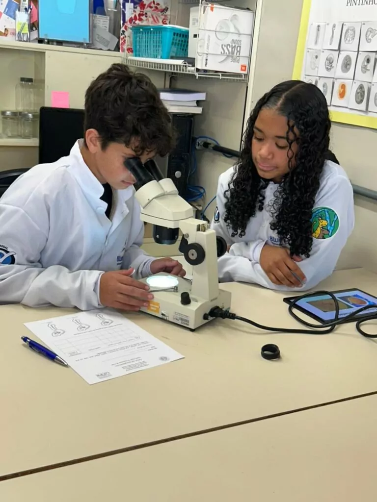A galeria de fotos mostra momentos da visita dos clubistas à UFPR. Trata-se de ambientes fechados como um auditório e um laboratório. Há fotos da palestra que mostra várias cadeiras enfileiradas ocupadas por estudantes e professores, que estão assistindo à palestrante e à projeção de slide sobre o Rio iguaçu. Nas fotos do laboratório, mostra pequenos grupos de estudantes ouvindo orientações dos professores, usando microscópios e tablets, fazendo anotações. Todos estão vestindo jalecos com expressões de concentração e interesse.