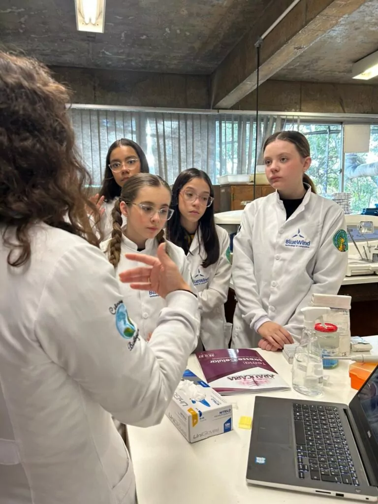 A galeria de fotos mostra momentos da visita dos clubistas à UFPR. Trata-se de ambientes fechados como um auditório e um laboratório. Há fotos da palestra que mostra várias cadeiras enfileiradas ocupadas por estudantes e professores, que estão assistindo à palestrante e à projeção de slide sobre o Rio iguaçu. Nas fotos do laboratório, mostra pequenos grupos de estudantes ouvindo orientações dos professores, usando microscópios e tablets, fazendo anotações. Todos estão vestindo jalecos com expressões de concentração e interesse.