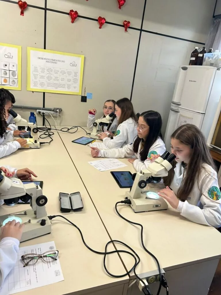 A galeria de fotos mostra momentos da visita dos clubistas à UFPR. Trata-se de ambientes fechados como um auditório e um laboratório. Há fotos da palestra que mostra várias cadeiras enfileiradas ocupadas por estudantes e professores, que estão assistindo à palestrante e à projeção de slide sobre o Rio iguaçu. Nas fotos do laboratório, mostra pequenos grupos de estudantes ouvindo orientações dos professores, usando microscópios e tablets, fazendo anotações. Todos estão vestindo jalecos com expressões de concentração e interesse.