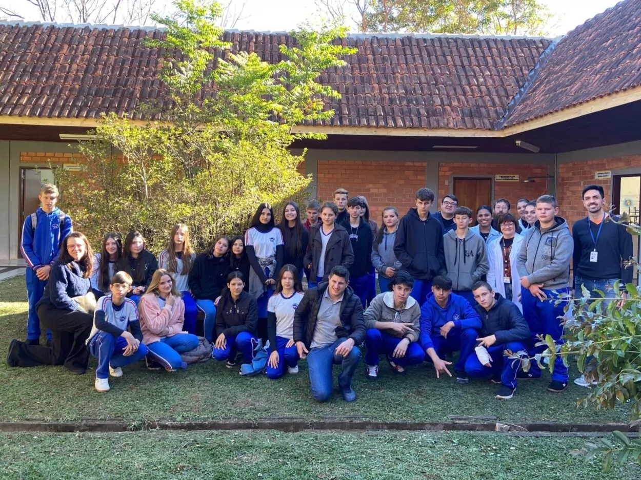 Foto de um grupo grande de estudantes e professores posando ao ar livre, em frente a um prédio com paredes de tijolos aparentes e telhado de cerâmica. Ao fundo, há vegetação e árvores. A maioria dos estudantes veste uniforme azul com detalhes brancos e vermelhos, e alguns usam roupas casuais. O grupo está dividido em duas fileiras: alguns agachados ou sentados na frente e outros em pé atrás. Todos olham para a câmera, muitos sorrindo. A luz do sol ilumina suavemente a cena.
