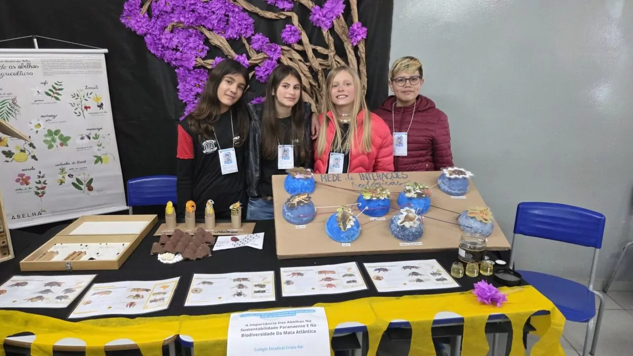 Quatro estudantes do Colégio Estadual Cristo Rei estão em pé atrás de uma mesa expositiva decorada com materiais didáticos sobre abelhas. São três meninas e uma menina. Eles usam crachás da Fieg e estão sorrindo. A mesa traz maquetes coloridas representando uma "rede de interações ecológicas", pequenos frascos com mel, cartazes informativos e modelos de abelhas. Ao fundo, há um mural com uma grande árvore feita de papel kraft com flores roxas em destaque. Um dos cartazes destaca a importância das abelhas na agricultura. A apresentação faz parte do projeto vencedor da Fieg na categoria Clubes de Ciências.