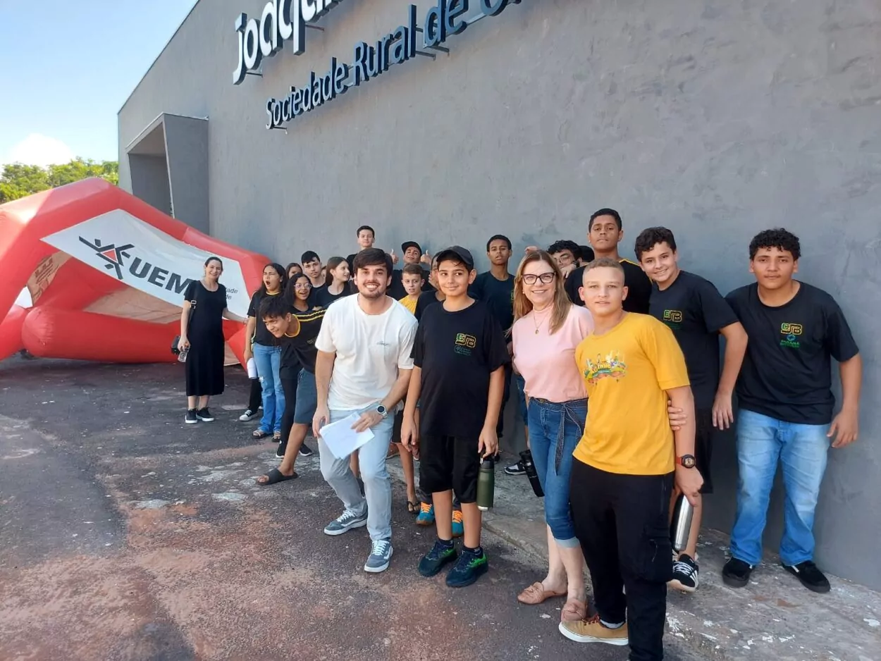 A foto é do Clube de Ciências Dengue Bot. A imagem mostra um grupo de pessoas, formado por adolescentes e alguns adultos, reunidos em frente a um prédio com a inscrição "Sociedade Rural de Umuarama". Eles estão participando de um evento. Muitos dos adolescentes estão usando uniformes escolares. Um dos adultos é o professor e está vestindo camiseta clara segurando algumas folhas. Há também uma mulher de óculos, com camiseta rosa, que aparenta ser uma das responsáveis pelo grupo. Ao fundo, vemos uma estrutura inflável vermelha e branca com o logo da UEM (Universidade Estadual de Maringá). O clima é descontraído e alegre, com o grupo reunido para a foto, muitos sorrindo e demonstrando bom humor.