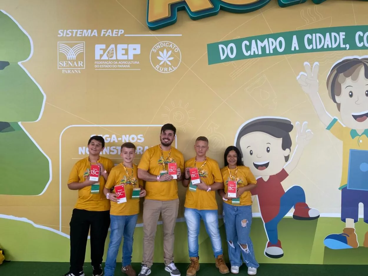 Esta imagem mostra um grupo de cinco pessoas, sendo quatro adolescentes e um adulto, todos posando para a foto em frente a um painel colorido e ilustrado. O painel tem os dizeres "Do Campo a Cidade, Conectando Saberes” e logotipos do Sistema FAEP, SENAR Paraná, FAEP (Federação da Agricultura do Estado do Paraná) e Sindicato Rural. Todos estão usando camisetas amarelas, provavelmente padronizadas para o evento, e crachás de identificação. Eles seguram um prêmio que é um celular. O ambiente é alegre e descontraído, com o painel de fundo ilustrado com personagens infantis, o que sugere que o evento tem caráter educativo e interativo, com foco em jovens.