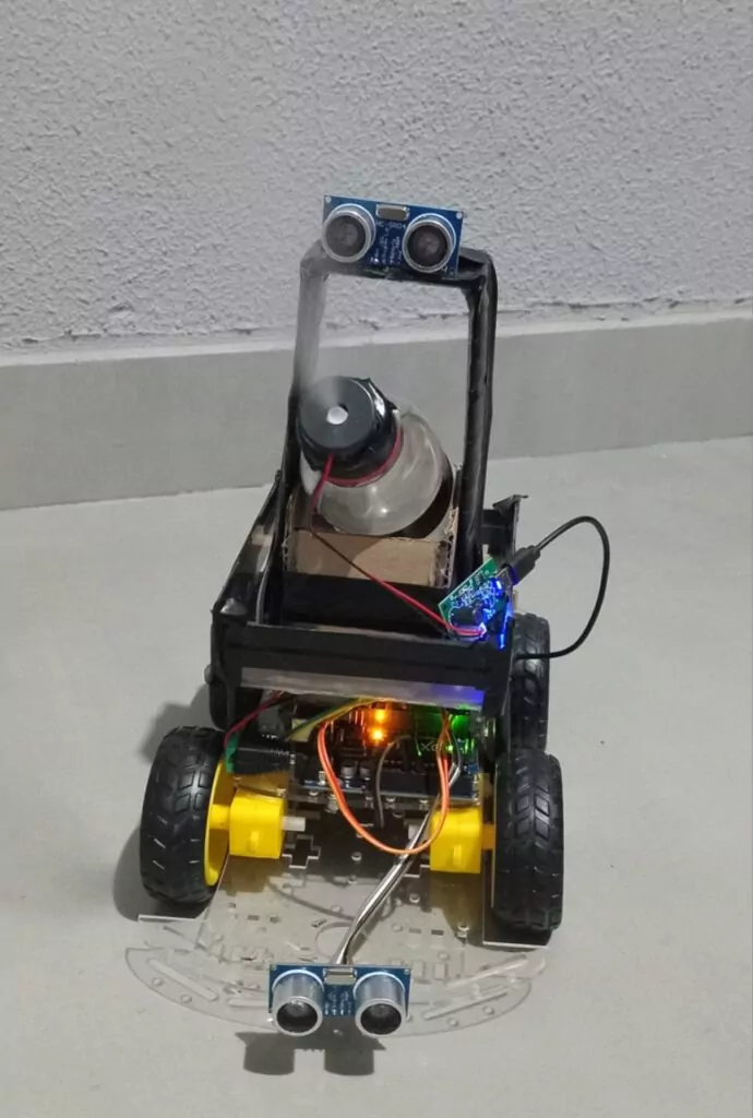 Esta imagem mostra um robô desenvolvido de forma artesanal. O robô possui uma estrutura montada com rodas, sensores ultrassônicos na parte frontal e superior, e componentes eletrônicos conectados, como placas de controle e fiação exposta. Na parte superior do robô, há um pequeno reservatório acoplado a um sistema que emite uma névoa, indicando a pulverização de um líquido, como parte da função do robô no combate à dengue. O sistema parece ter sido projetado para se locomover de maneira autônoma ou controlada, detectando obstáculos através dos sensores e liberando o produto quando necessário, visando eliminar ou repelir o mosquito Aedes aegypti. Esse tipo de projeto integra tecnologia e soluções sustentáveis para problemas reais, como o combate à dengue.
