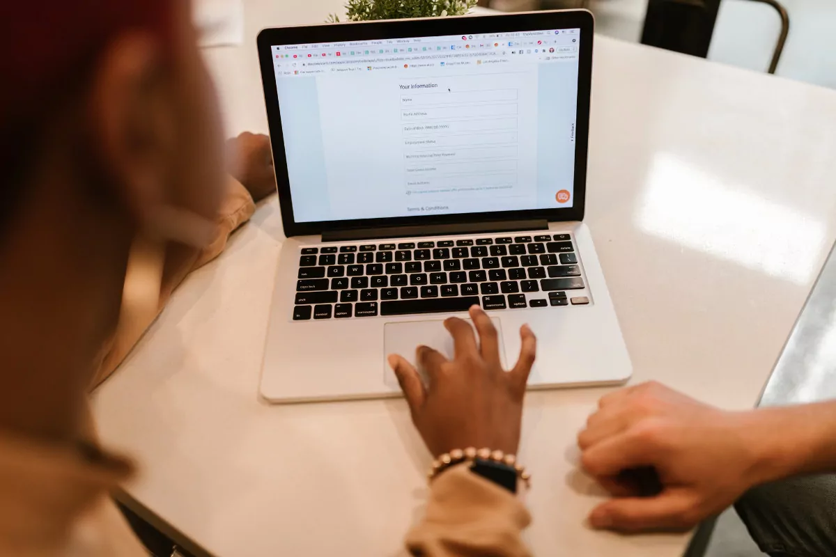 A imagem mostra duas pessoas, que estão sentadas lado a lado em uma mesa branca, olhando para a tela de um notebook prateado com teclado preto. Na tela do notebook, há um formulário digital aberto, com o título “Your Information” ("Suas Informações", em inglês). Abaixo do título, há campos a serem preenchidos. No fundo da imagem, há uma pequena planta no centro da mesa. A iluminação é clara, com reflexos de luz branca na superfície brilhante da mesa.