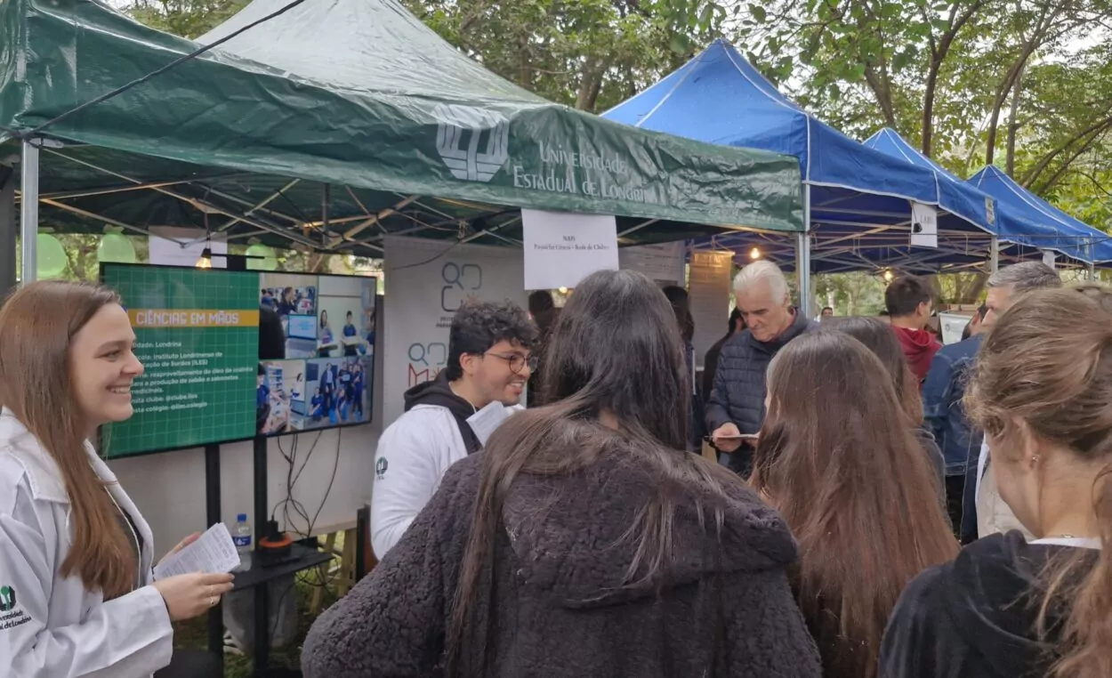 A imagem mostra representantes do NAPI Paraná Faz Ciência apresentando a Rede de Clubes para os visitantes da 13° Feira das Profissões da Universidade Estadual de Londrina. A equipe está posicionada em um estande e o público é formado em sua maior parte por estudantes de escolas da região. Os alunos prestam atenção na explicação.