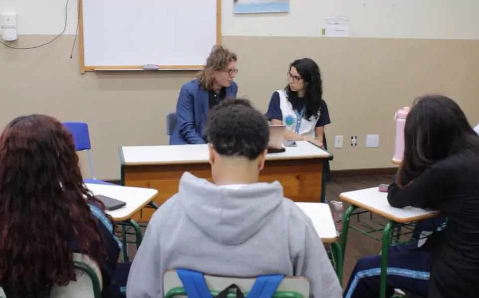 Na foto, vemos um momento de conversa atenta entre o cineasta, que tem cabelos compridos e óculos, vestido com blazer azul, e a professora, vestida com jaleco branco e logo da escola. Ambos estão sentados à frente da sala de aula, atrás de uma mesa, olhando-se nos olhos em um diálogo concentrado. Sobre a mesa, há um notebook fechado. Ao redor deles, estudantes ocupam cadeiras escolares, formando uma roda. A maioria está voltada para o centro, prestando atenção. O ambiente da sala é simples, com paredes em dois tons, um quadro branco ao fundo e uma pintura de paisagem logo acima.