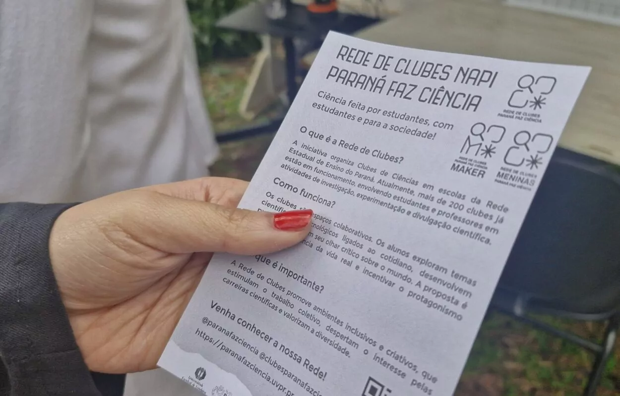 A imagem mostra uma mulher segurando um dos panfletos que foram distribuídos pela equipe NAPI UEL durante a Feira das Profissões. O material de divulgação indica os objetivos da Rede de Clubes e seu papel na sociedade, além de conter um QR Code para que os visitantes possam acessar as redes sociais dos clubes de ciencias da região.