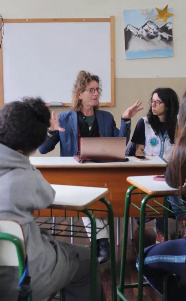 A foto mostra uma sala de aula onde o cineasta Rudolf Mestdagh está sentado atrás de uma mesa, conversando com o grupo de estudantes do clube. Essa pessoa está ao centro, utiliza um notebook aberto à sua frente e gesticula enquanto fala. Ao lado, a professora que rege o clube ouve com atenção. Os estudantes estão sentados em carteiras escolares, prestando atenção e, em alguns casos, fazendo anotações. Ao fundo, há um quadro branco, uma pintura com montanhas e estrelas douradas coladas na parede. O ambiente é simples e típico de uma sala de aula, com paredes em dois tons e móveis escolares padrão.