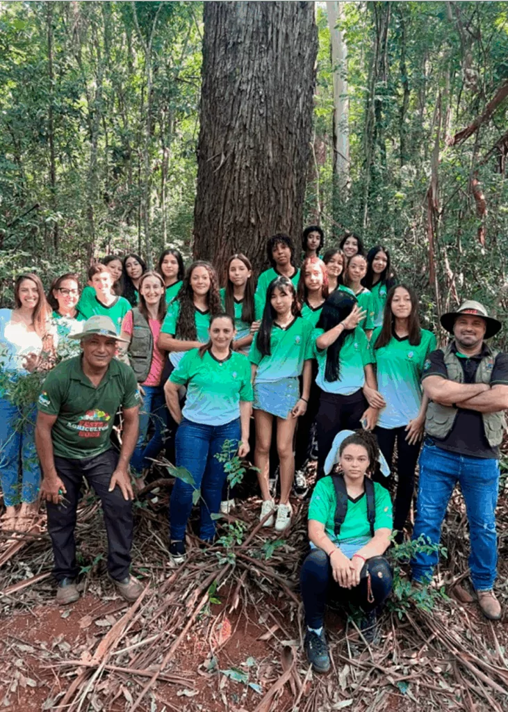 Na imagem, alunos e professores do Clube de Ciências do Colégio Estadual Vital Brasil participam de uma atividade ao ar livre, na trilha ecológica. A turma, vestindo camisetas verdes do uniforme escolar, posou ao redor de uma grande árvore, símbolo da importância da preservação ambiental. A saída de campo faz parte das ações do clube, que estimula a observação, o contato com o meio ambiente e o aprendizado prático sobre ecologia e sustentabilidade.