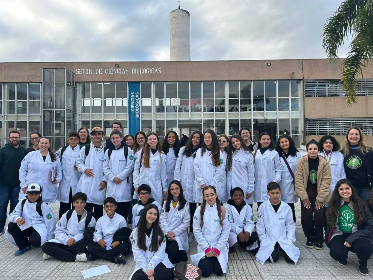 A foto mostra um grande grupo de estudantes e professores posando para uma foto em frente a um prédio institucional chamado "Setor de Ciências Biológicas". O grupo está reunido ao ar livre, em um dia nublado. A fachada do prédio é de cor clara, com janelas de vidro amplas, e ao centro há uma faixa azul vertical com os dizeres “Ciências Biológicas”. No topo da construção, ergue-se uma torre cilíndrica branca. A maioria das pessoas na foto está vestindo jalecos brancos. Muitos dos jalecos possuem o logotipo da instituição, o brasão da escola ou outro emblema no braço. Entre os participantes, há adolescentes, pré-adolescentes e alguns adultos, que são professores. Algumas pessoas estão agachadas à frente para que todos caibam bem na foto. Há uma palmeira visível no canto direito da imagem, e o chão é pavimentado com pequenos blocos claros. O clima parece frio, pois algumas pessoas estão de casaco ou com as mãos nos bolsos. Todos estão sorrindo ou com expressões alegres, transmitindo um sentimento de união e entusiasmo.