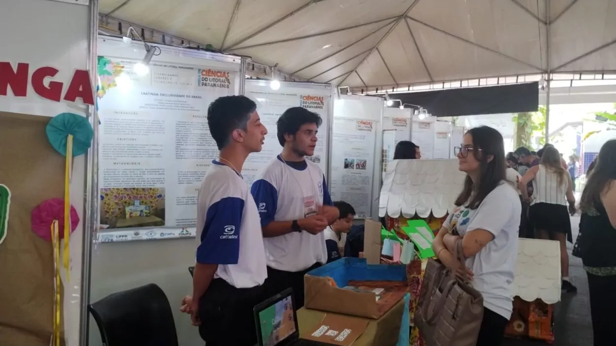 Na foto, dois estudantes, usando camisetas brancas com mangas azuis, apresentam seu projeto sobre a Caatinga para uma visitante, que escuta atentamente com os braços cruzados e segura uma bolsa bege. Ao fundo, vê-se um painel expositivo com o título “Caatinga: Exclusividade do Brasil” e outros trabalhos da feira de ciências, todos sob uma grande tenda. Sobre a mesa, há uma maquete colorida e um notebook aberto, compondo a exposição do grupo.