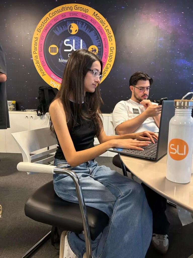 Laura (16 anos), foi uma das alunas selecionadas no Paraná, pelo Colégio CEEP Pedro Boaretto Neto, de Cascavel, no Sustainable Living Innovators (SLI). Ela integrou a equipe que embarcou do Brasil até outro continente para uma aventura científica em Portugal. Na foto é possível vê-la sentada em frente ao computador, discutindo com outro integrante sobre o tema de trabalho da equipe.