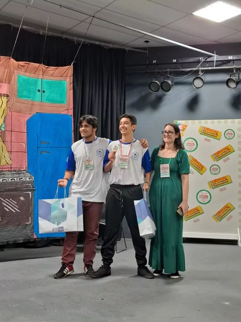 A imagem mostra dois estudantes e com a professora Andreia Petruy em um palco durante um evento. Os dois jovens, usando camiseta branca com mangas azuis, estão sorrindo, cada um segurando uma sacola de premiação. Um deles exibe também uma medalha no peito. Ao lado deles, a professora, de vestido verde, sorri enquanto posa para a foto. Ao fundo, há um painel colorido com a logo da "Ciência do Litoral Paranaense" e um cenário decorativo que imita uma cozinha desenhada. O clima é de celebração e reconhecimento.