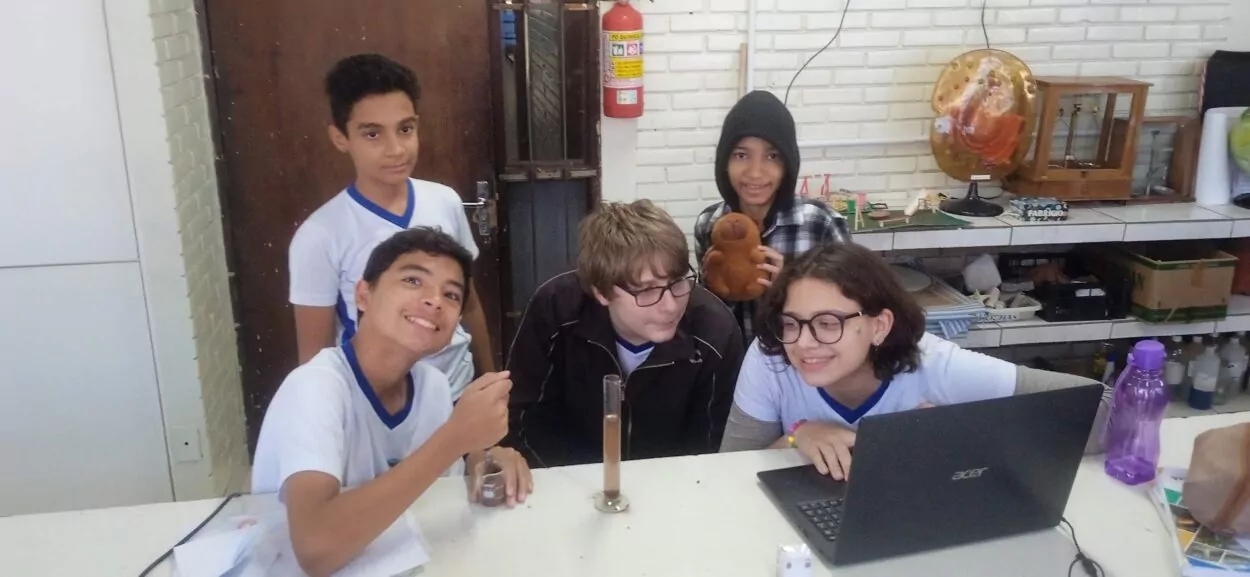 A imagem mostra um grupo de cinco estudantes em um ambiente escolar. Eles estão reunidos ao redor de uma mesa branca que contém um notebook, um cilindro medidor com líquido marrom, um dado, uma garrafa roxa de água e alguns papéis. Os estudantes vestem uniformes escolares com camiseta branca de gola azul, exceto um deles, que usa um casaco preto. Eles parecem estar engajados em uma atividade prática ou experimento científico, e demonstram expressões de curiosidade e entusiasmo. Ao fundo, há uma parede de tijolos brancos, estantes com diversos materiais didáticos e uma maquete. Um dos estudantes segura uma pelúcia marrom e está usando um moletom com capuz. Também é possível ver um extintor de incêndio preso à parede e diversos objetos organizados nas prateleiras, indicando um ambiente de aprendizado.