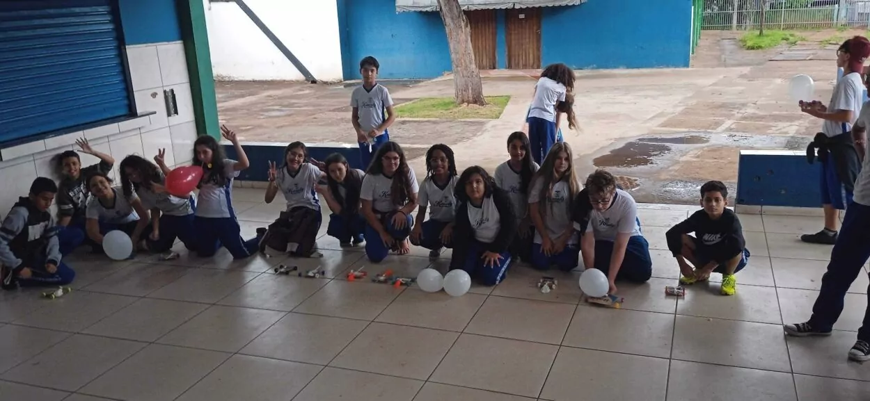 A imagem mostra um grupo grande de estudantes reunidos em um espaço coberto da escola, provavelmente participando de uma atividade prática. A maioria dos alunos está usando uniforme escolar, composto por camiseta branca com detalhes azuis e calça azul. Eles estão agachados ou sentados no chão, lado a lado, formando uma longa fileira. À frente de cada grupo de alunos, há carrinhos artesanais feitos com materiais simples e recicláveis (como palitos, tampinhas e bexigas), sugerindo uma atividade de construção de carrinhos movidos a ar (balões inflados). Alguns alunos seguram ou preparam balões para os carrinhos, demonstrando entusiasmo e concentração. Ao fundo, observa-se parte do pátio da escola com piso de cimento, árvores e muros azuis. Também há alguns estudantes em pé. A atmosfera é descontraída, colaborativa e educativa.