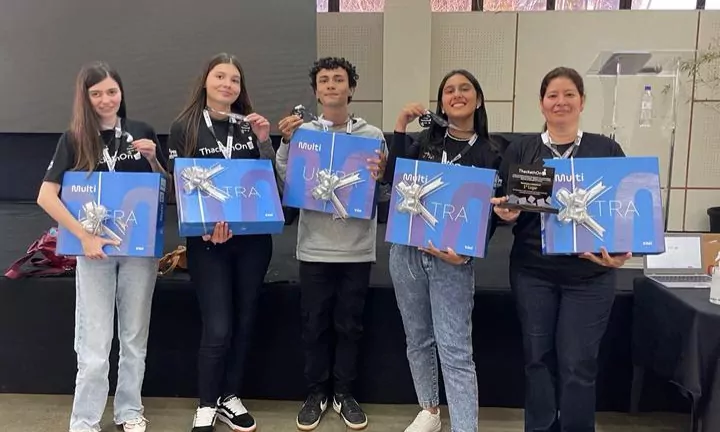 A imagem mostra um grupo de cinco pessoas (quatro jovens e uma mulher adulta) em pé, sorrindo e segurando prêmios. Todos usam camisetas pretas do evento "ThackathOn". Cada pessoa segura uma grande caixa azul com a palavra "ULTRA" em destaque e a marca "Multilaser", envolvida com um laço prateado, sugerindo que seja um notebook. Além disso, cada um segura um pequeno troféu preto.