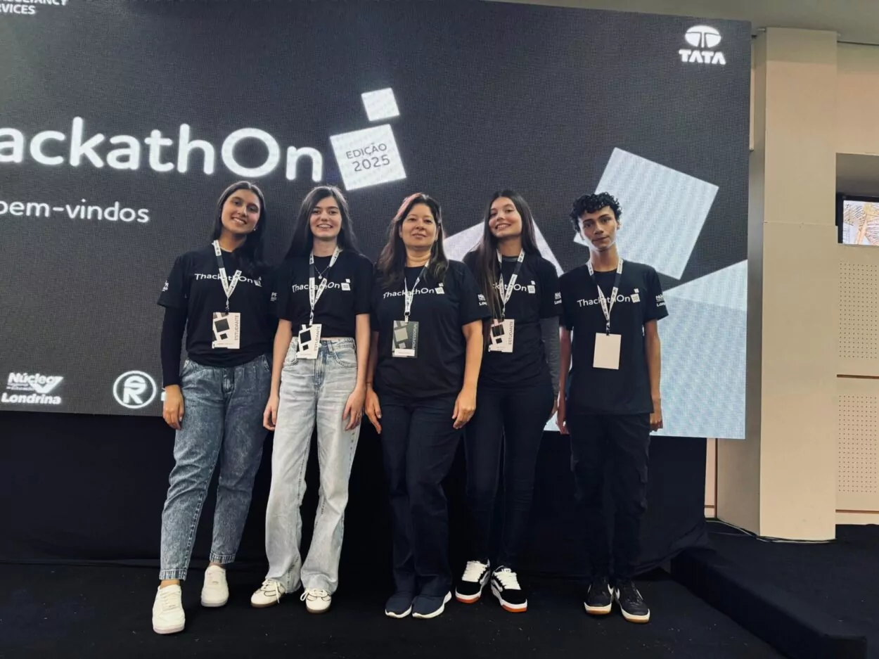 A imagem mostra um grupo de cinco pessoas (quatro jovens e uma mulher adulta) em pé sobre um palco, lado a lado, sorrindo para a câmera. Todos estão usando camisetas pretas com o logotipo “ThackathOn Edição 2025” estampado na frente, além de crachás pendurados no pescoço. Eles estão posicionados em frente a um telão digital grande que exibe o nome do evento em letras brancas. As pessoas na imagem são as vencedoras do evento.