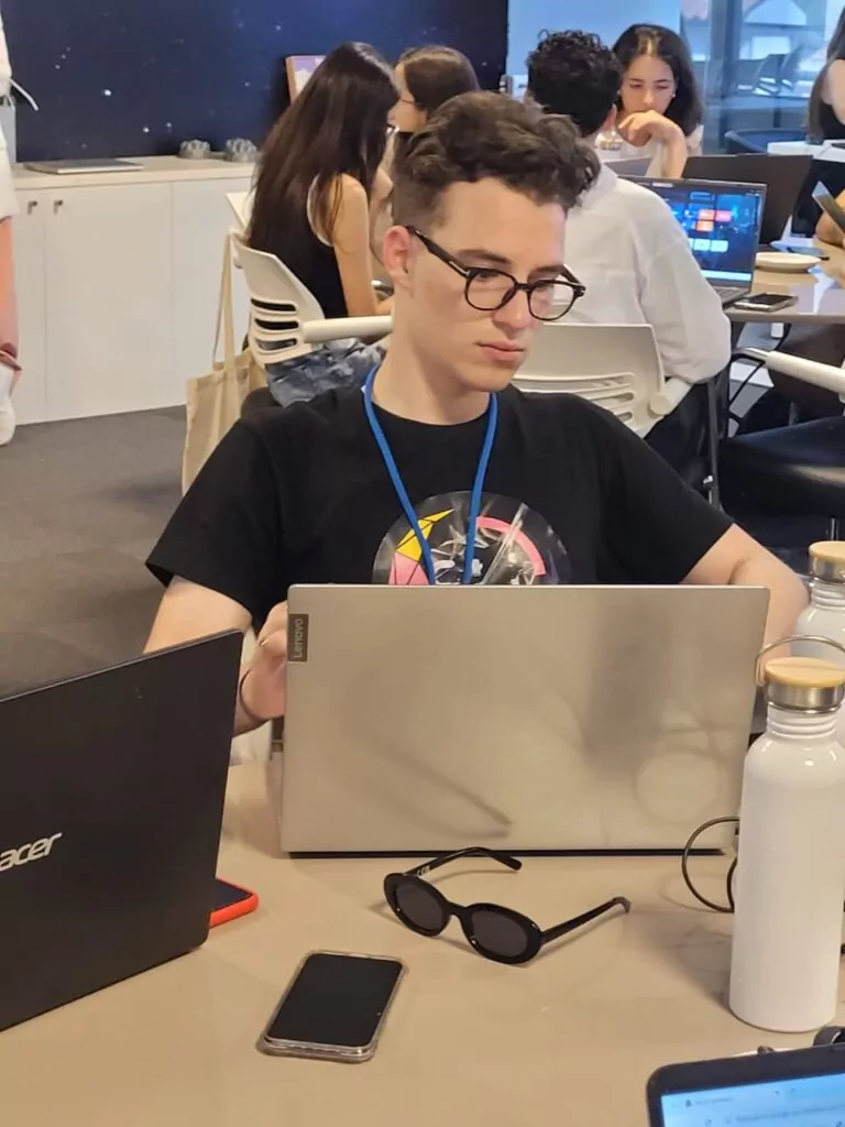 Um estudante está sentado em uma mesa de trabalho coletiva, utilizando um notebook. Ele veste camiseta preta estampada e crachá azul, com óculos de grau de armação escura. À sua frente, sobre a mesa, há outro notebook fechado, um celular, um par de óculos de sol e duas garrafas brancas de metal. Ao fundo, outros estudantes trabalham em laptops em um ambiente moderno, com paredes decoradas por imagens que remetem ao espaço. A cena registra um momento de concentração durante atividades acadêmicas em grupo.