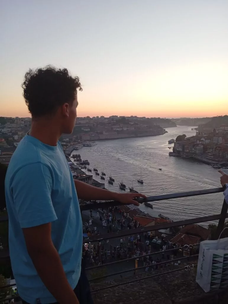 A imagem mostra um jovem de camiseta azul clara, visto de perfil, observando o pôr do sol sobre um rio. Ele está encostado em um corrimão, no alto de um mirante, com vista para a cidade e para diversas embarcações atracadas ou navegando. Ao fundo, é possível ver prédios, pontes e o céu em tons de laranja e rosa, refletindo na água. Na parte inferior da cena, há um grande grupo de pessoas reunidas, possivelmente turistas, aproveitando a vista. A atmosfera transmite tranquilidade e contemplação.