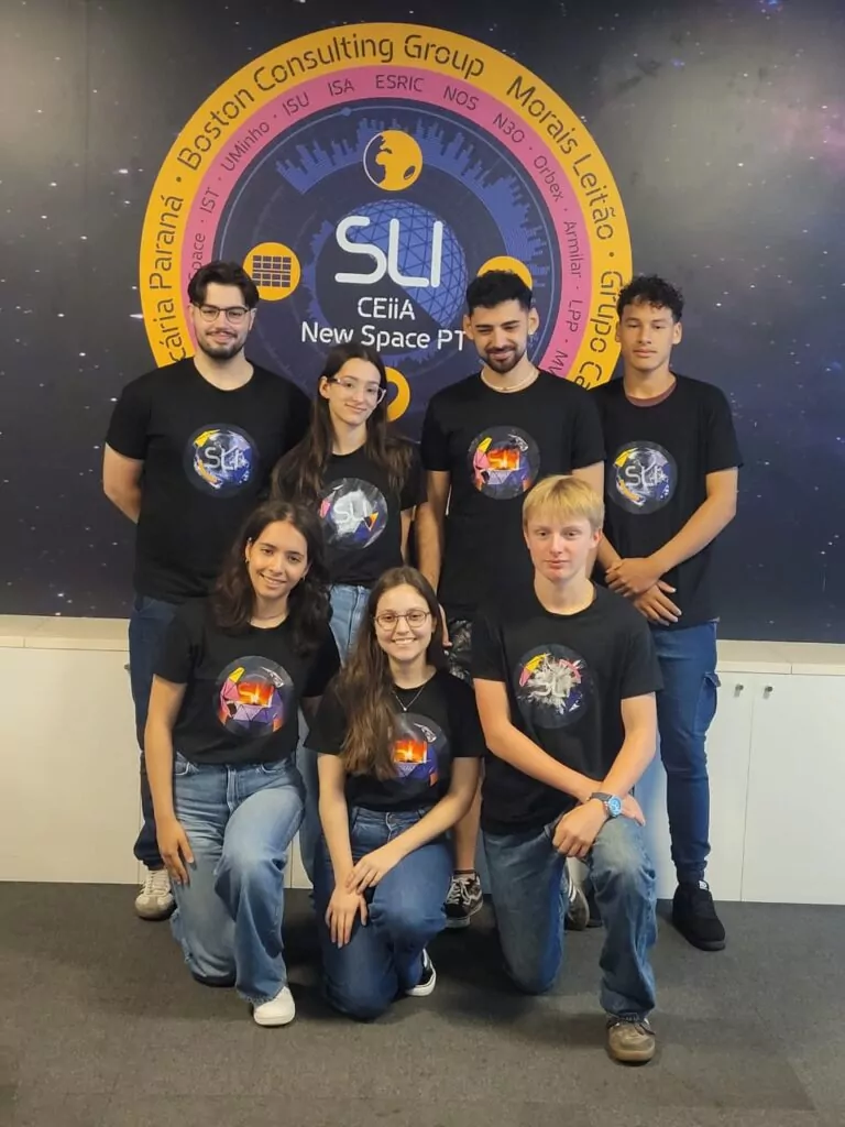 A imagem mostra um grupo de sete jovens posando para a foto em um ambiente interno. Eles vestem camisetas pretas com estampas coloridas relacionadas ao espaço e ao evento SLI (Sustainable Living Innovators), e usam calças jeans ou escuras. Ao fundo, há um grande painel com o logotipo do SLI e do CEiiA New Space PT, cercado por nomes de instituições e empresas parceiras. O grupo está disposto em duas fileiras: quatro pessoas em pé na parte de trás e três agachadas ou ajoelhadas na frente, todas sorrindo levemente para a câmera.