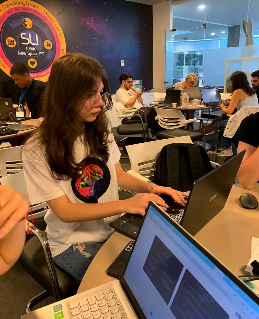 A fotografia mostra uma jovem de cabelos longos e castanhos, usando óculos e camiseta branca com estampa colorida, sentada em uma mesa redonda e trabalhando em um notebook preto. À frente dela, há outro notebook aberto com uma tela clara. No fundo, outras pessoas também trabalham em computadores. Na parede, há um grande painel com o logotipo do programa “SLI – CEiiA New Space PT”, em um fundo que lembra o espaço sideral, com estrelas e planetas.
