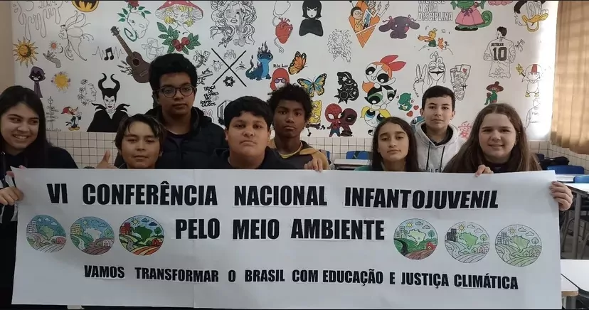 Oito estudantes posam segurando uma faixa branca com os dizeres: “VI Conferência Nacional Infantojuvenil pelo Meio Ambiente – Vamos transformar o Brasil com Educação e Justiça Climática”. Ao fundo, há uma parede repleta de desenhos coloridos feitos à mão, com elementos da cultura pop, flores, animais e frases diversas. A sala é iluminada por luz natural que entra pela janela à direita da imagem. Os alunos e alunas estão sorrindo levemente e demonstram orgulho por participarem da conferência.