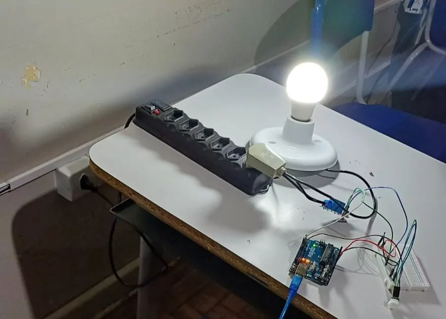 A imagem mostra o sensor de movimento construído pelos estudantes. Há uma lâmpada acesa, ligada em uma tomada. Há fios que conectam o equipamento a outros materiais. Ele foi montado com kits de robótica.
