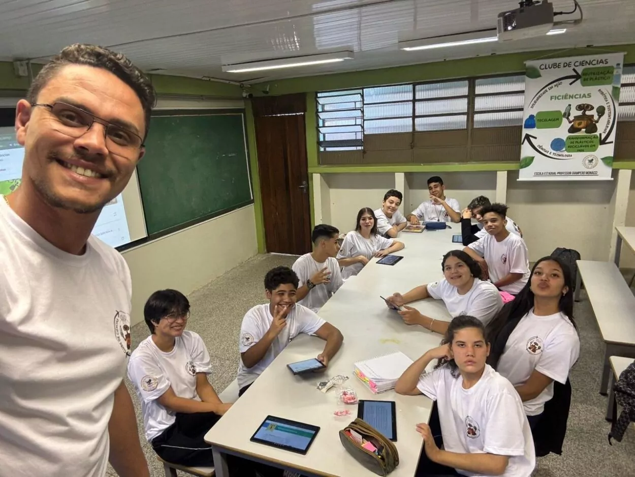 A primeira fotografia é do Clube Jovens Cientistas Kennedy. A imagem mostra um grupo grande de estudantes posando para a foto em um pátio externo, com árvores de folhagem vermelha ao fundo e um muro branco atrás deles. A maioria dos alunos veste uniforme escolar. Algumas alunas estão agachadas na frente, junto com duas mulheres adultas, enquanto o restante da turma permanece em pé. Um homem adulto está à esquerda do grupo. O clima da foto é de descontração e união. A segunda fotografia é do Clube Robótica e Sustentabilidade, Solidariedade em Ação. A imagem mostra uma sala de aula onde um grupo de estudantes, todos usando camiseta do uniforme escolar, está reunido em torno de uma mesa retangular. Alguns alunos estão com tablets e cadernos, demonstrando estarem em atividade de estudo. No canto esquerdo, em primeiro plano, um professor sorri para a câmera enquanto tira a selfie com a turma ao fundo. Na parede, há um cartaz do Clube de Ciências, que traz ilustrações relacionadas à reciclagem, reutilização de plástico e reaproveitamento de materiais. O ambiente é bem iluminado, com quadro verde e janelas amplas.