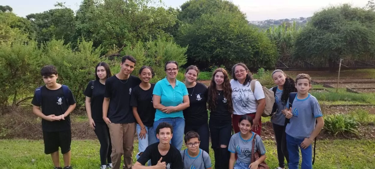 A primeira imagem é do Clube Experimentando a Ciência: Descobrindo cores e novos sabores. A imagem mostra um grupo de estudantes e professores posando para uma foto ao ar livre, em frente a uma placa de madeira com o desenho de uma abelha e a palavra “Meliponário”. O local é arborizado, com gramado verde, árvores grandes e, ao fundo, um prédio de tijolos com telhado de telhas vermelhas. A maioria dos alunos veste uniforme escolar, alguns usam mochilas. Estão sorridentes e organizados ao redor da placa. A segunda imagem é do Clube Ilha da Ciência. A imagem mostra um grupo de estudantes e duas mulheres posando em um ambiente ao ar livre, com árvores, arbustos e canteiros de horta ao fundo. A maioria dos alunos veste camisetas com logotipo escolar, e alguns carregam mochilas. O grupo está sorridente e reunido de forma descontraída, alguns em pé e outros agachados na frente.