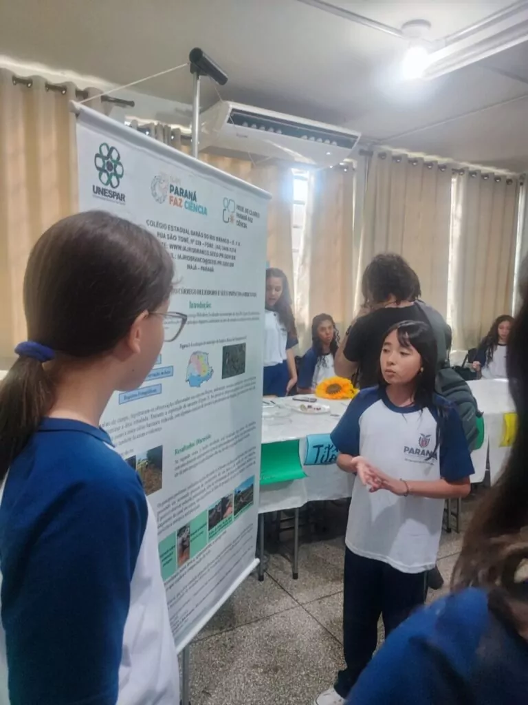 Na foto, uma estudante apresenta seu trabalho durante a 1ª Feira de Ciências da Unespar – Campus Paranavaí. Ela veste o uniforme escolar e está em pé ao lado de um banner científico que exibe o título do projeto, desenvolvido pelo Colégio Estadual Barão do Rio Branco, em Ivaiporã/PR, vinculado à Rede de Clubes Paraná Faz Ciência.