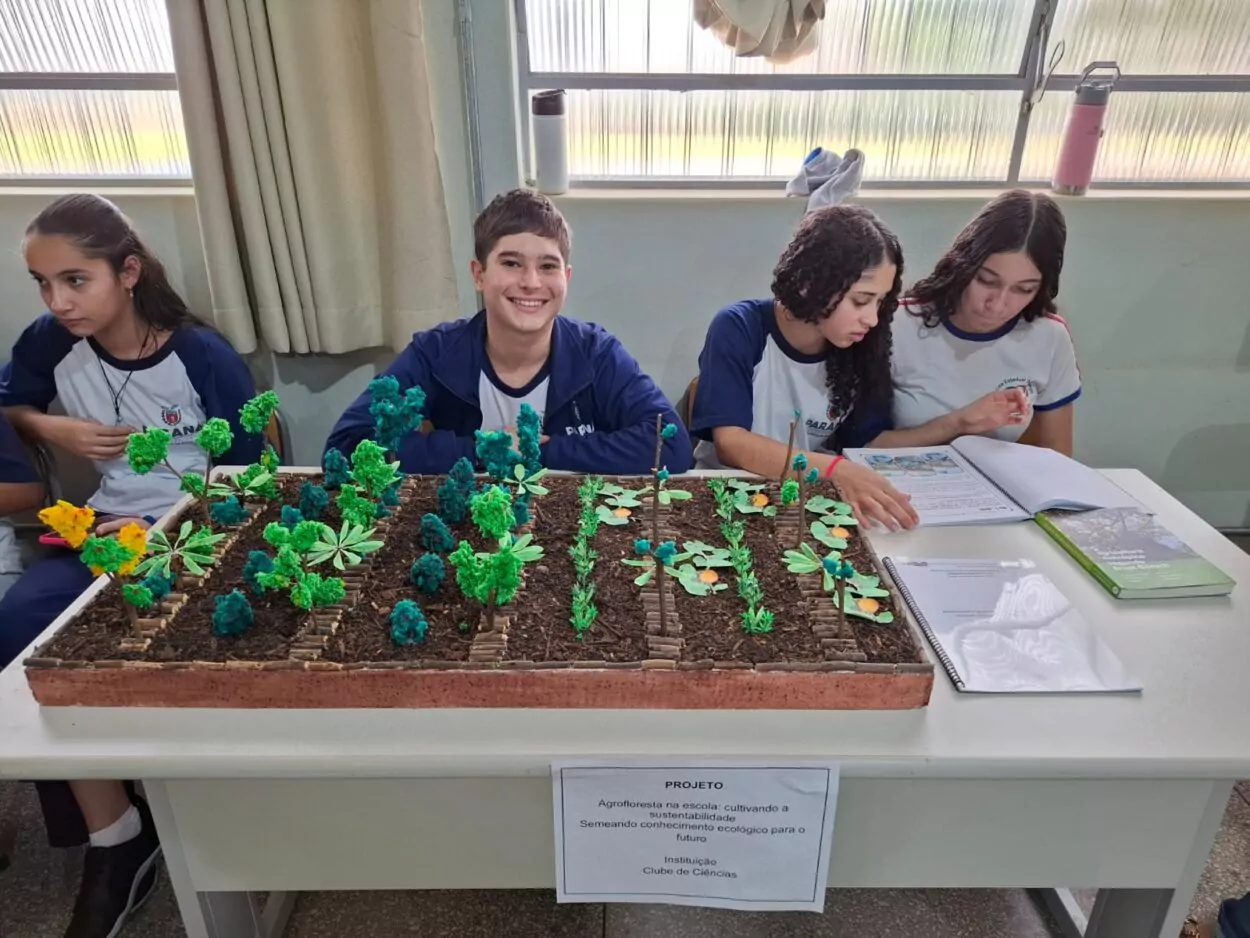 Quatro estudantes estão sentados lado a lado em uma mesa, vestindo uniformes escolares. À frente deles, sobre a mesa, há uma maquete representando uma agrofloresta, feita com materiais que simulam árvores, plantas e canteiros. O trabalho está identificado por uma placa que traz o título: “Agrofloresta na escola: cultivando a sustentabilidade e semeando o conhecimento ecológico para o futuro”, desenvolvido por um Clube de Ciências.