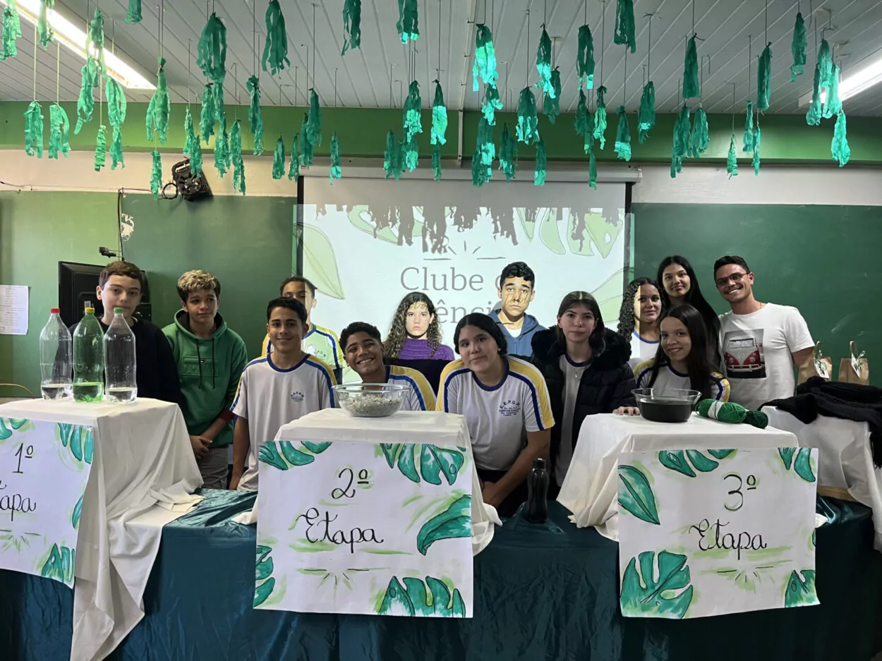 Foto dos clubistas e do professor coordenador do Clube ‘Robótica e Sustentabilidade, Solidariedade em Ação’ no dia da apresentação da culminância do primeiro semestre de 2025. A imagem mostra um grupo de estudantes posando juntos em uma sala de aula decorada. No teto, há tiras de papel verde penduradas, simulando folhas ou plantas, criando um clima de natureza. Ao fundo, um projetor exibe a frase “Clube de Ciências”, acompanhada de ilustrações de pessoas. Na frente, três mesas estão cobertas com toalhas brancas e têm cartazes escritos “1ª Etapa”, “2ª Etapa” e “3ª Etapa”, decorados com desenhos de folhas verdes. Sobre as mesas há materiais usados em experimentos, como garrafas plásticas - PET, uma bacia com flakes, um pote com corante, fios de lã, touca e cachecol e outros objetos. Os estudantes sorriem e posam para a foto, transmitindo um clima de trabalho em grupo e entusiasmo pelas atividades do clube de ciências.