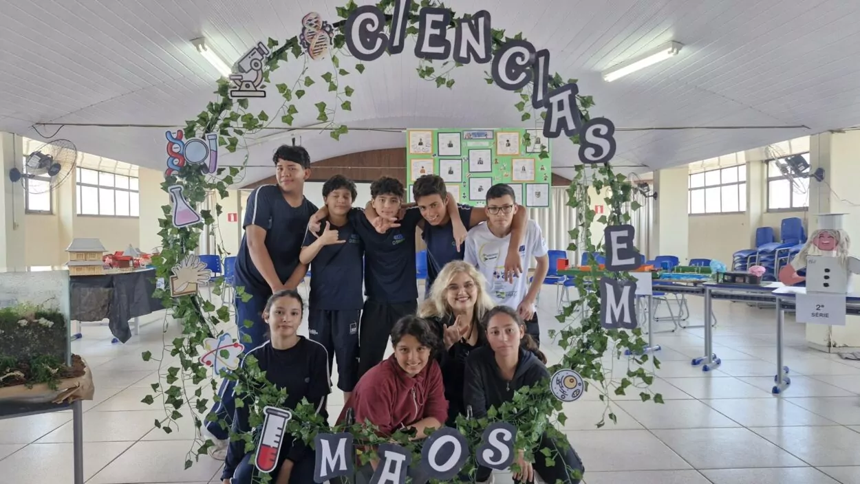Estudantes e professora sorrindo dentro de um arco feito com folhas artificiais e ilustrações de elementos científicos, como tubos de ensaio, DNA e microscópio. No arco, está escrito “CIÊNCIAS EM MÃOS”