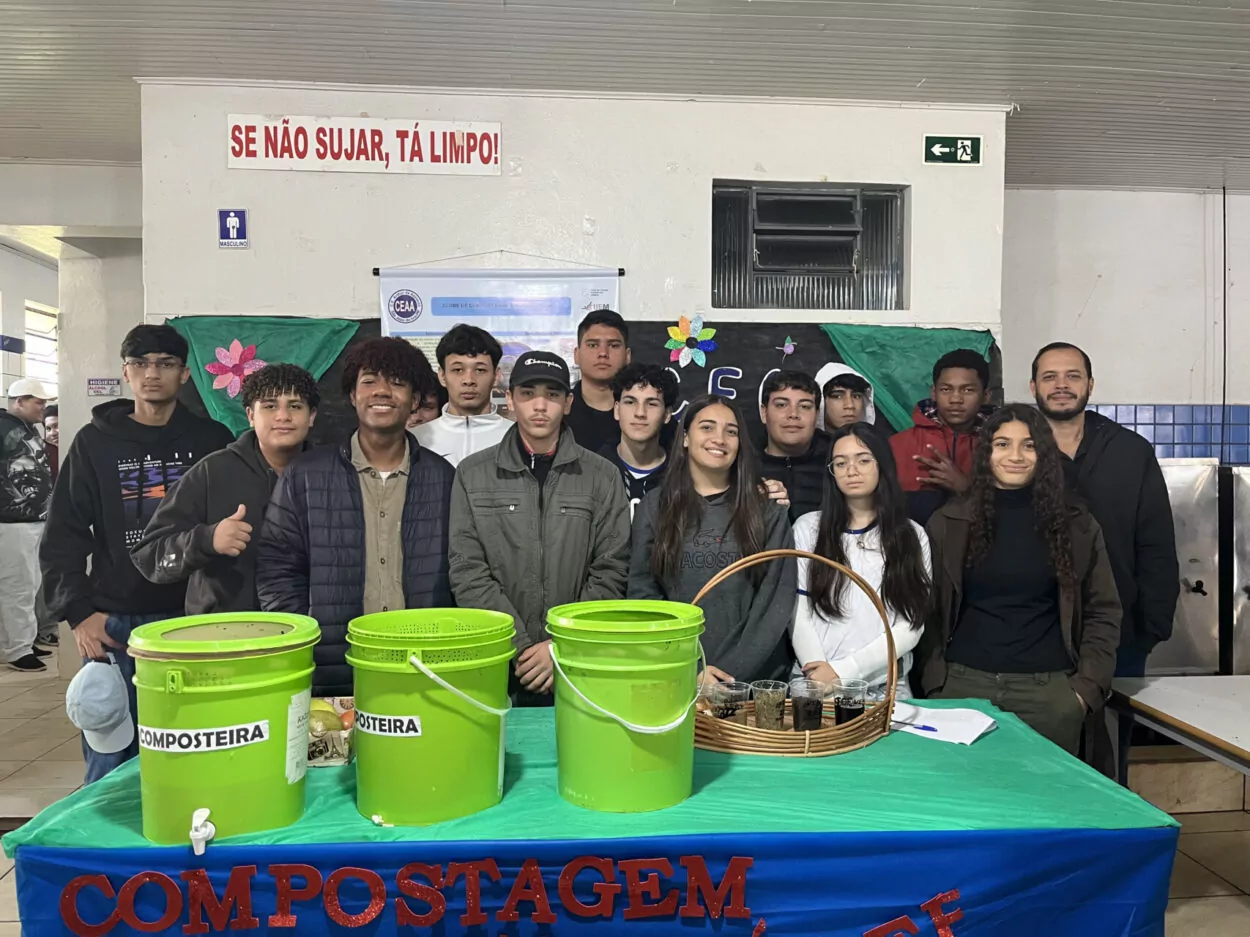 A imagem é do clube “Horta e Compostagem Escolar” do Colégio Estadual Arthur de Azevedo em São João do Ivaí. Na fotografia é possível ver um grupo de estudantes e um professor posando para a foto em um ambiente escolar. Eles estão atrás de uma mesa coberta com um tecido azul e verde, onde estão dispostos três baldes verdes rotulados como “Composteira” e um cesto com recipientes contendo composto. Ao fundo, há um painel decorado com flores coloridas e o nome do clube. O clima é de apresentação sobre compostagem e sustentabilidade.