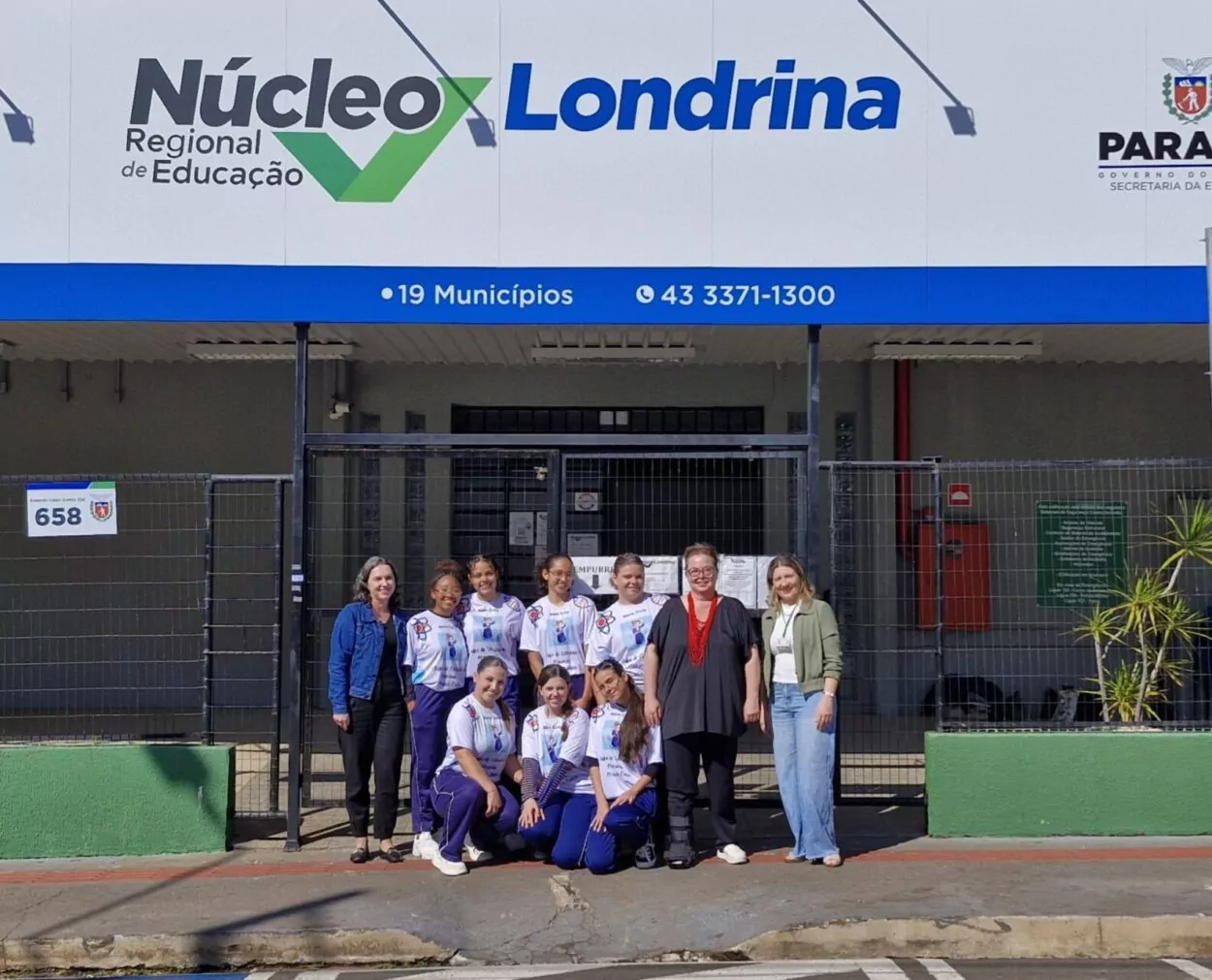 Um grupo de pessoas está posando para a foto do lado de fora, em frente a um prédio com a placa “Núcleo Regional de Educação Londrina – Paraná”. As estudantes continuam com seus uniformes e as três mulheres adultas estão juntas, sorrindo. O grupo está atrás de um portão de grade preta, em calçada de rua. O sol ilumina bem a cena.