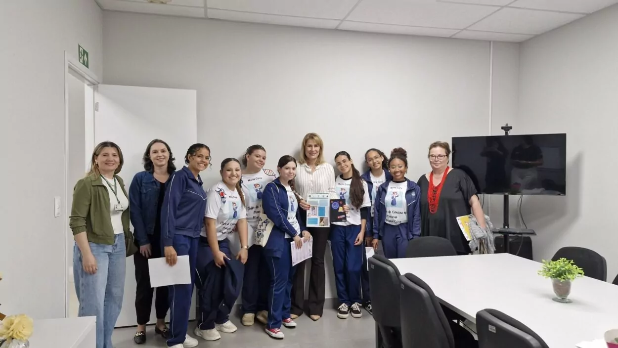 Algumas estudantes estão sentadas ao redor de uma mesa redonda branca. Elas usam o mesmo uniforme de Marie Curie e seguram papéis coloridos. Na mesa, há folhetos impressos em tons de roxo e branco, com textos sobre ciência e empoderamento feminino.