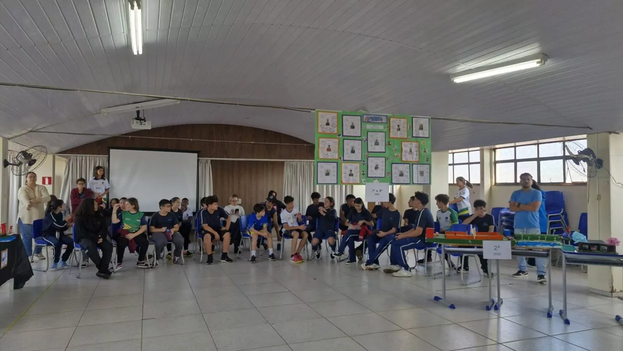 Apresentação do projeto do Clube “Ciência em Mãos” em um amplo salão escolar, bem iluminado, com piso claro, teto de forro branco e janelas grandes ao fundo. Ao centro da imagem, diversos alunos estão sentados em cadeiras organizadas em semicírculo. Na parede central, há um painel com cartazes contendo trabalhos escolares. No canto direito da imagem, há mesas com maquetes representando projetos. Adultos observam e acompanham o evento dos cantos da sala.