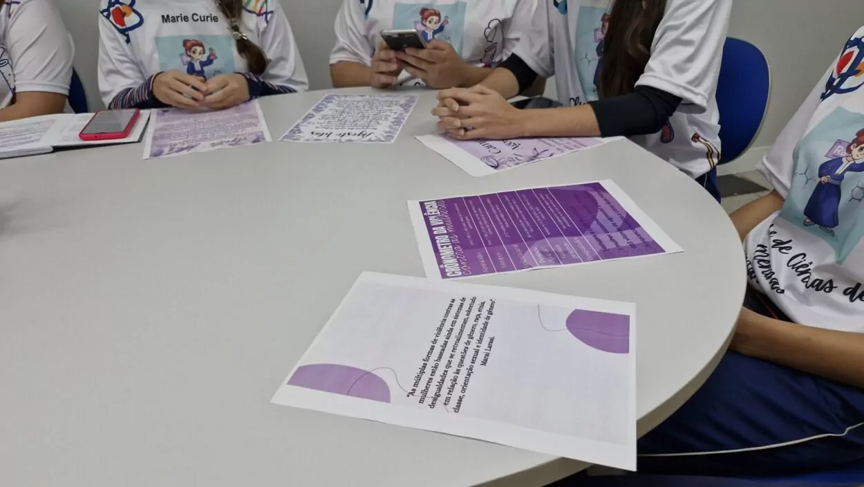 Algumas estudantes estão sentadas ao redor de uma mesa redonda branca. Elas usam o mesmo uniforme de Marie Curie e seguram papéis coloridos. Na mesa, há folhetos impressos em tons de roxo e branco, com textos sobre ciência e empoderamento feminino.